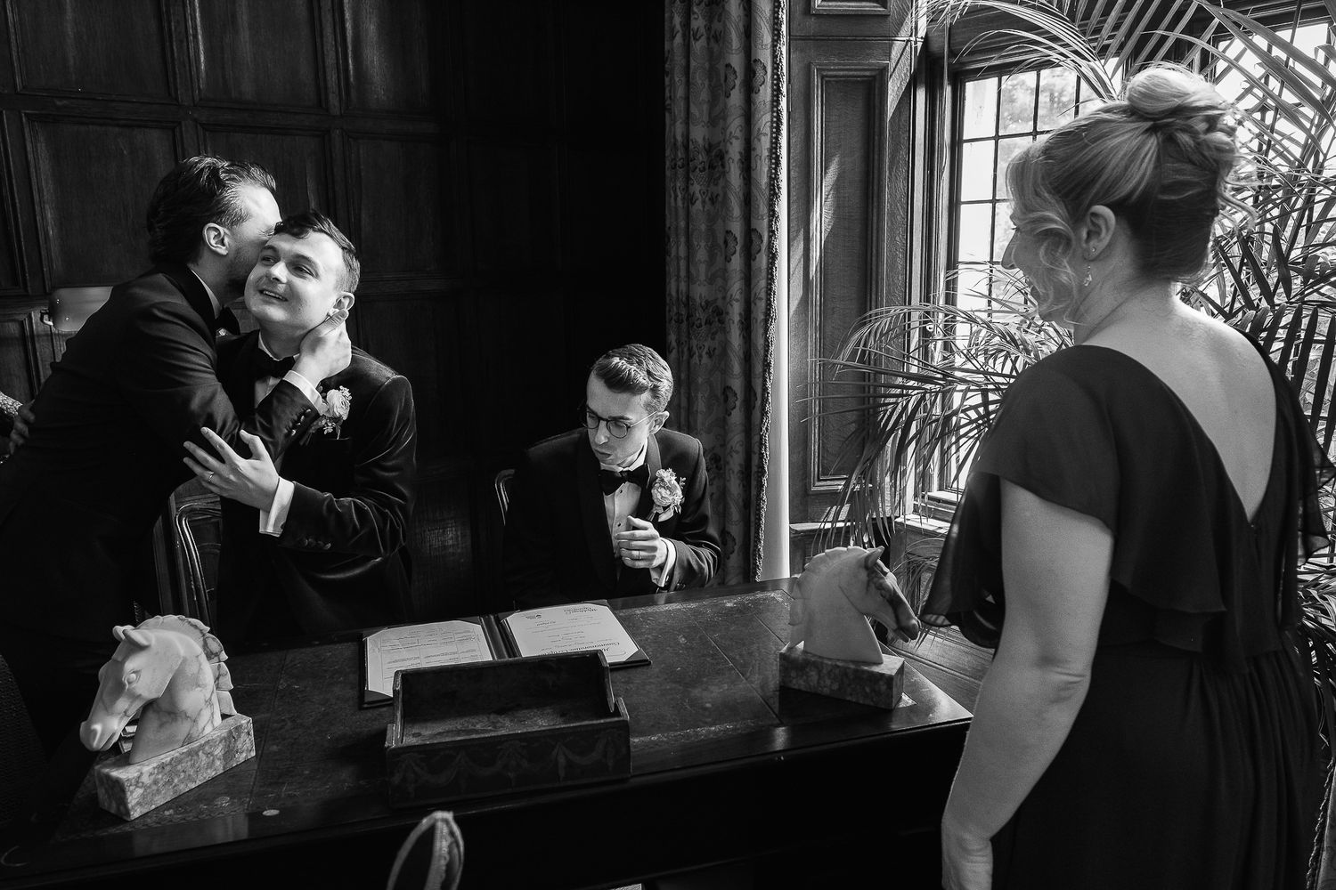 Black and white wedding photo of grooms embracing near a desk while guests look on in an elegant room.