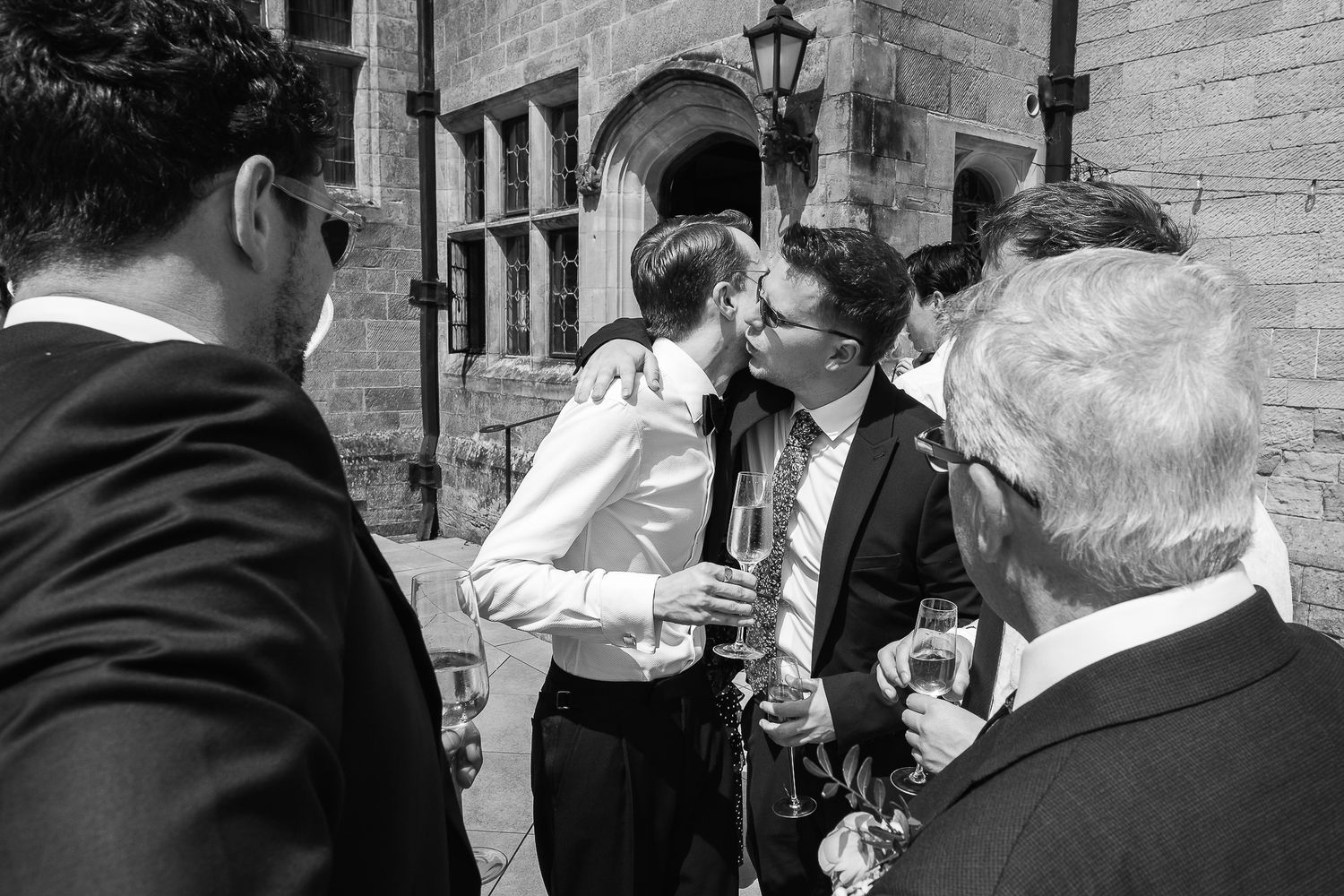 Black and white wedding photo of groom being congratulated by guests outside a historic stone building.