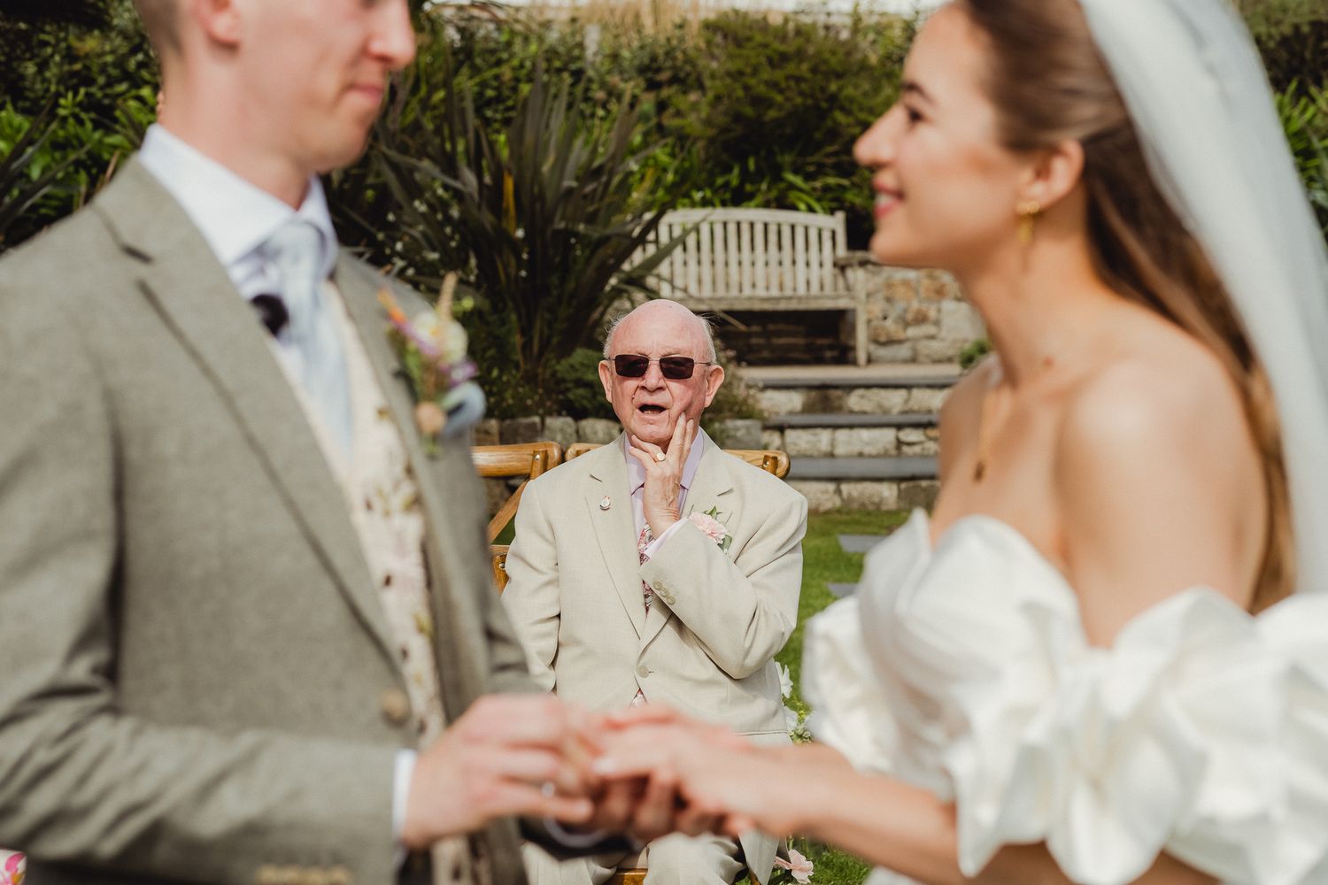 A candid wedding ceremony moment shows a guest reacting with joy in the background as a couple exchanges rings outdoors.