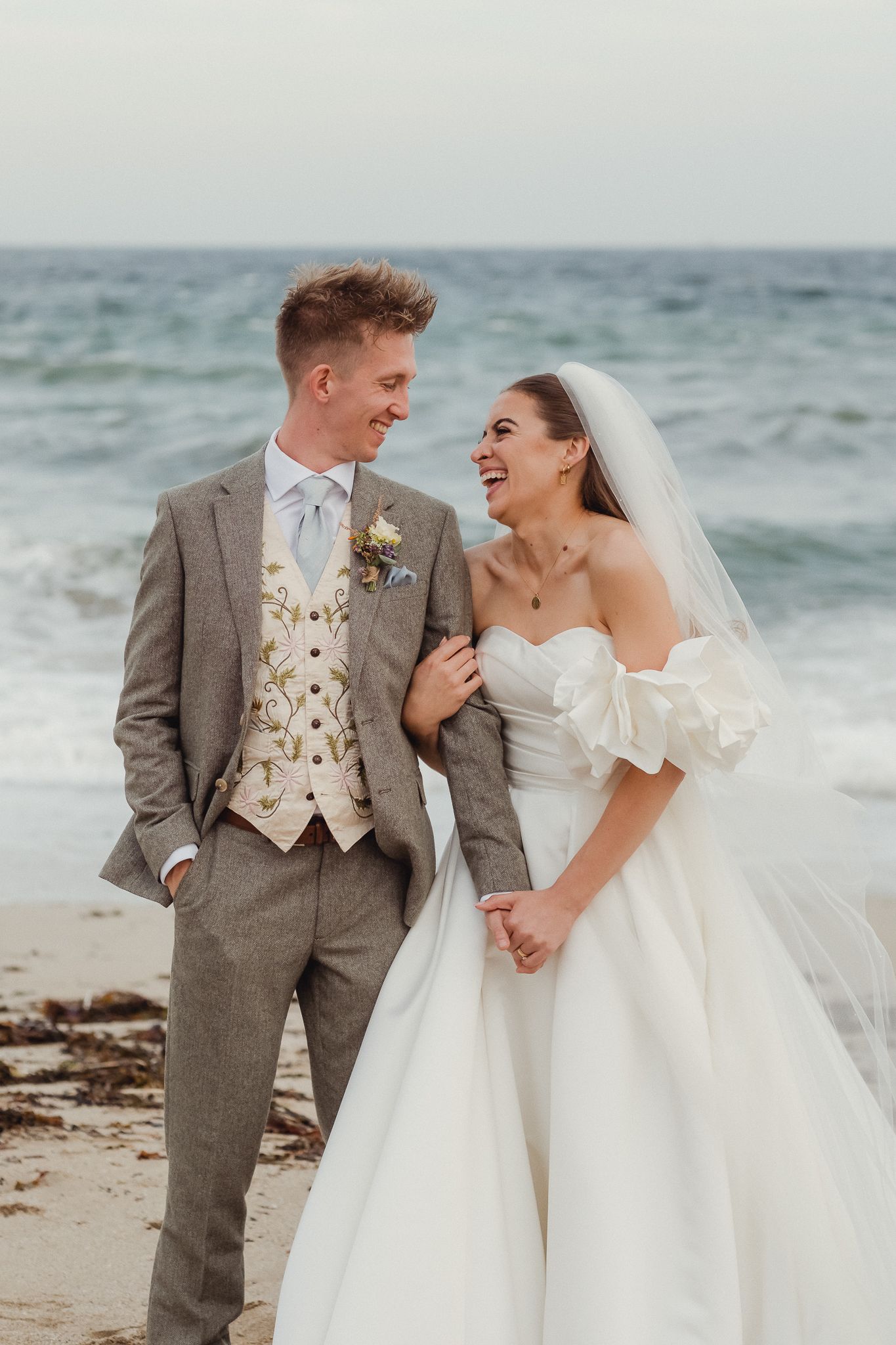 A joyful wedding couple shares a romantic moment on a cloudy beach with waves crashing in the background.