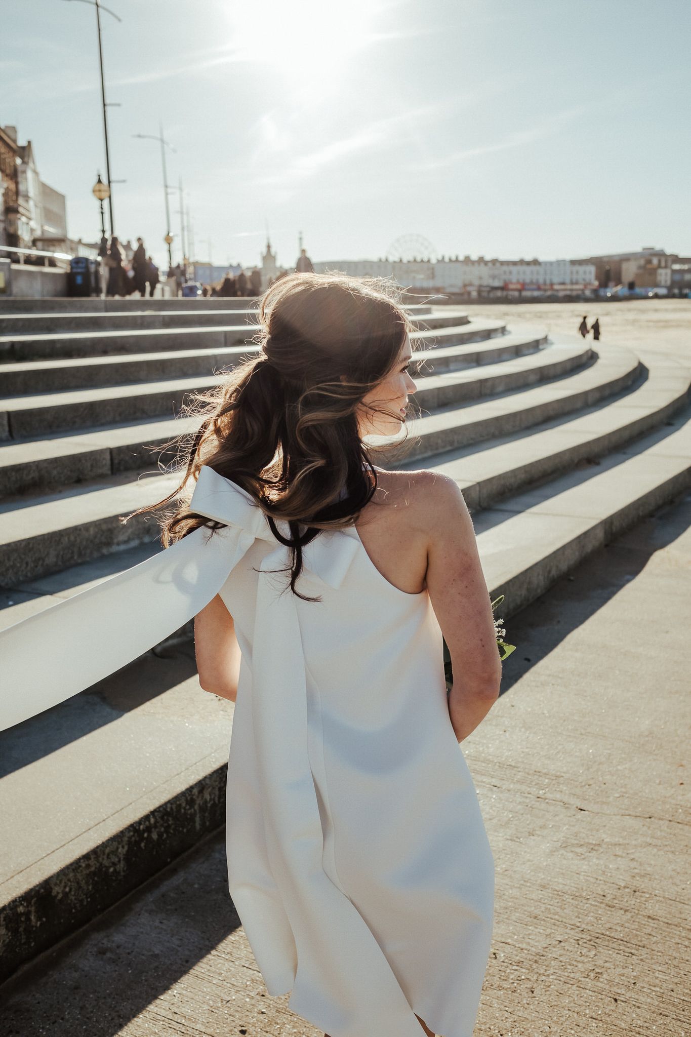 A woman in a white top sits on geometric concrete steps on a sunny day as her hair blows in the wind.
