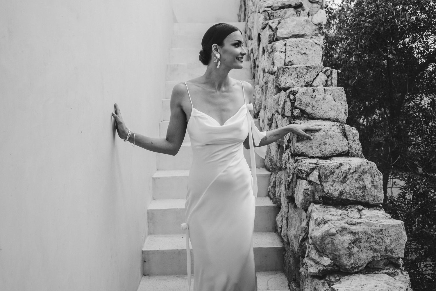 Black and white photo of a bride in a sleek white gown posing elegantly against a stone wall and staircase.
