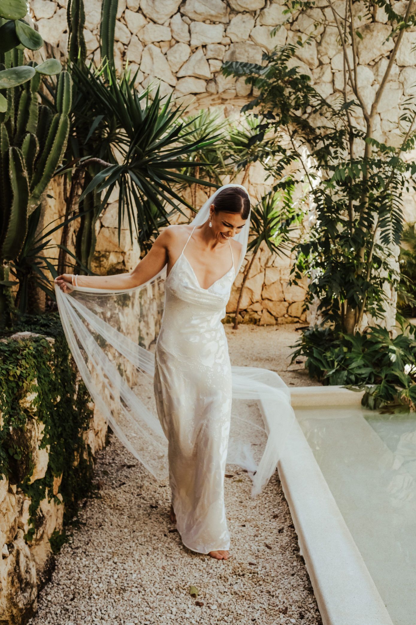 Elegant white wedding dress flowing in the breeze against stone wall and tropical plants in outdoor garden setting.