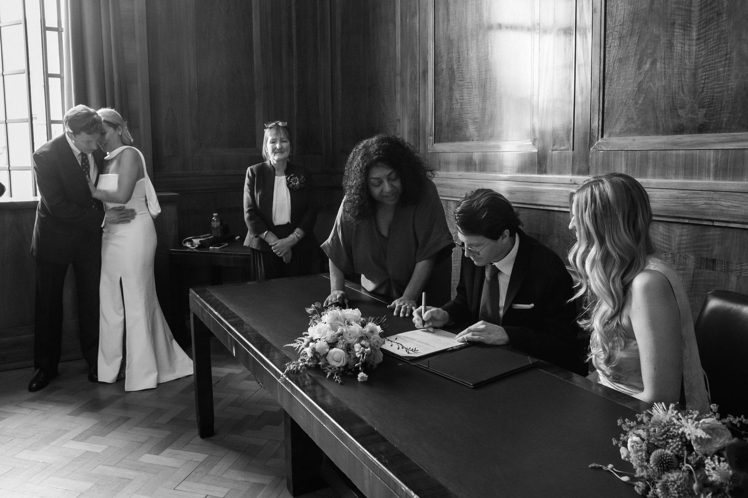 Couple signs wedding register at ornate wooden table with officiant and witnesses in black and white.
