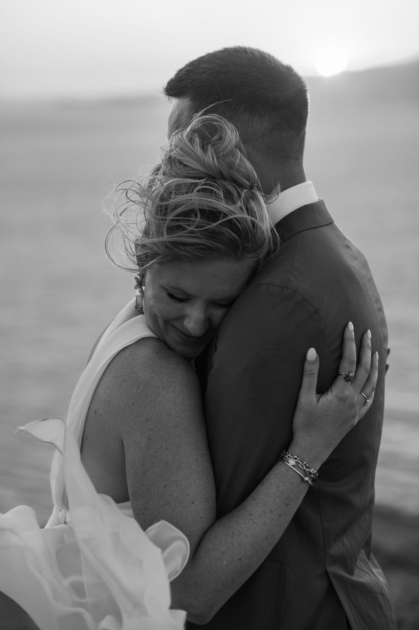 Black and white photo of a bride and groom embracing at sunset, the bride smiling with wind-blown hair.