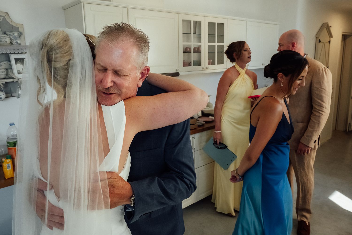 Bride and father share emotional hug before wedding ceremony while guests mingle in background.