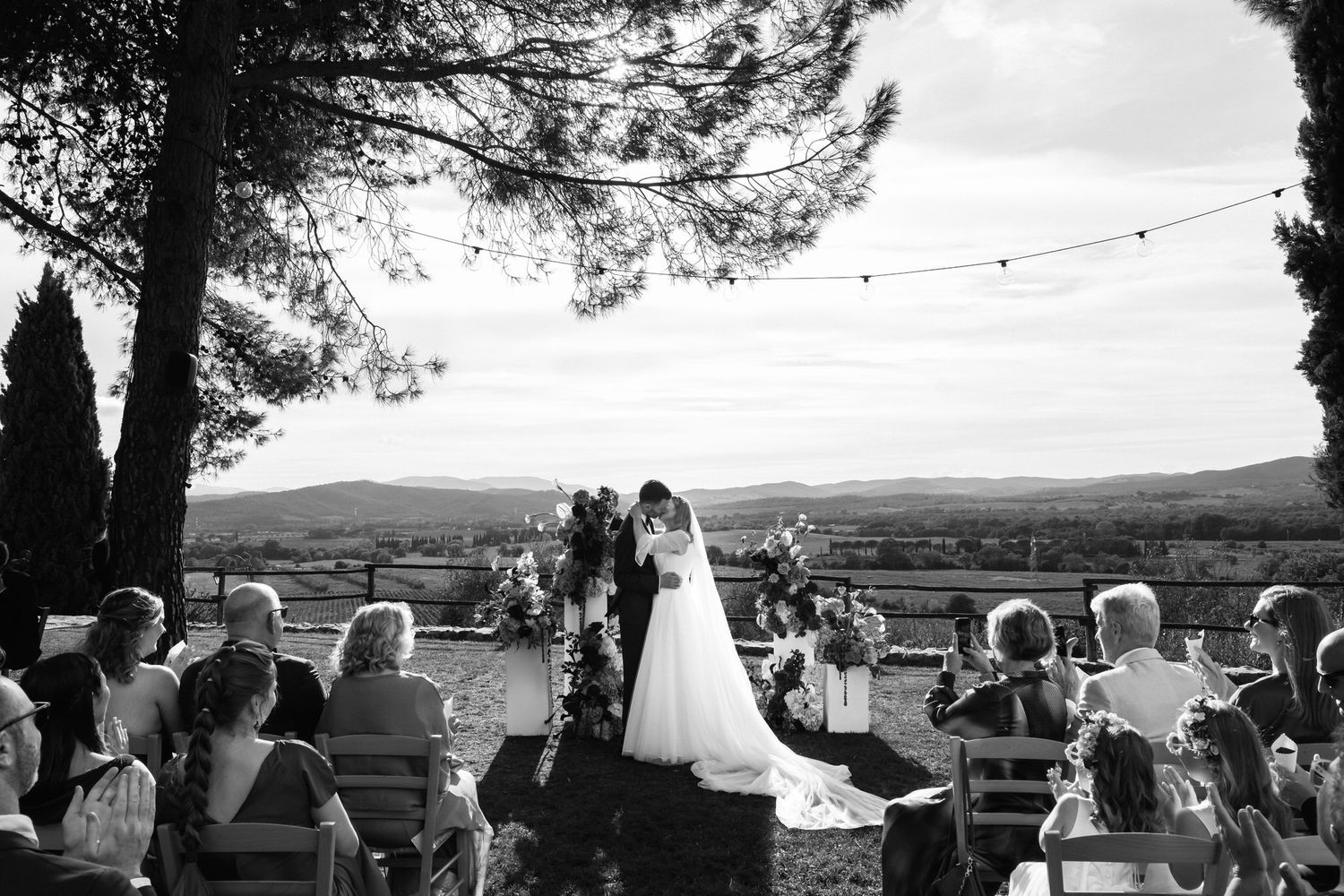 Black and white photo of a couple kissing at an outdoor hilltop wedding ceremony with seated guests, string lights and rolling hills behind.