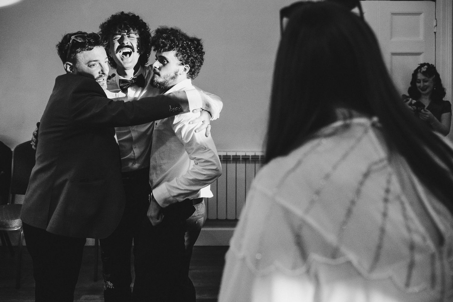 Black and white photo of three men in suits laughing and embracing in a joyful group hug at a celebration while guests look on.
