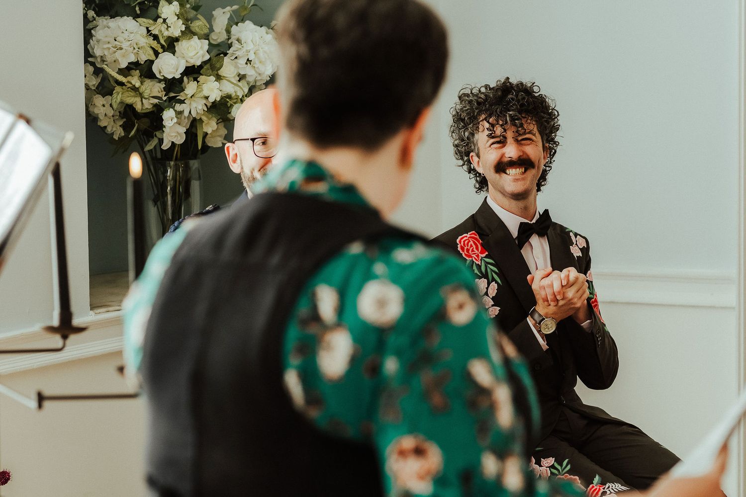 Smiling groom with curly hair and a mustache clasps his hands in a dark suit with a red boutonniere during an intimate wedding ceremony.
