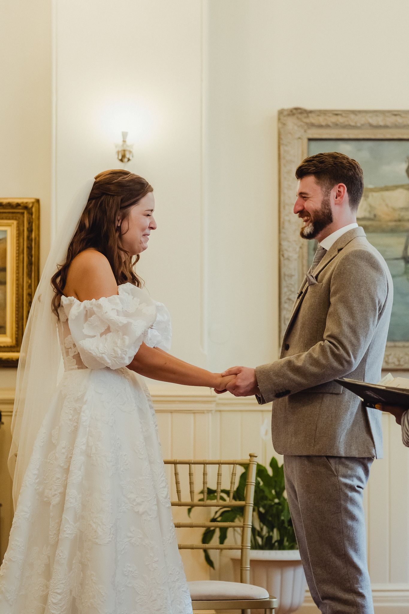 A couple shares a joyful moment during their wedding ceremony in an elegant room with framed artwork and soft lighting.