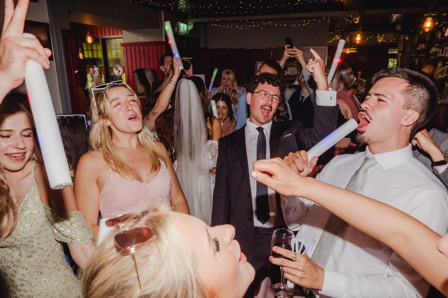 Joyful wedding celebration with people dancing and having fun under ambient lighting at a reception venue.