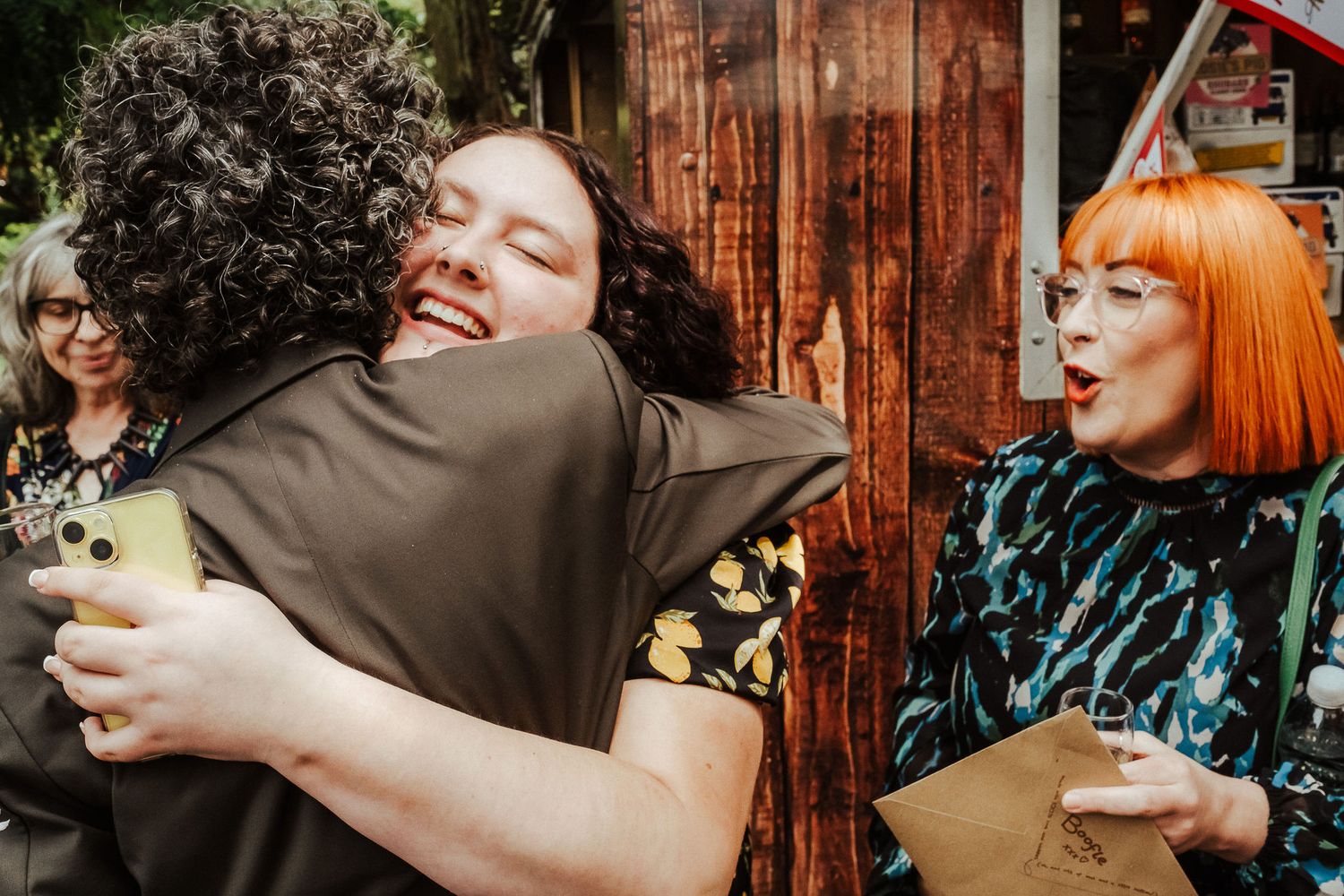 Two people share a joyful hug outdoors while a smiling red-haired woman in glasses looks on beside a rustic wooden wall.