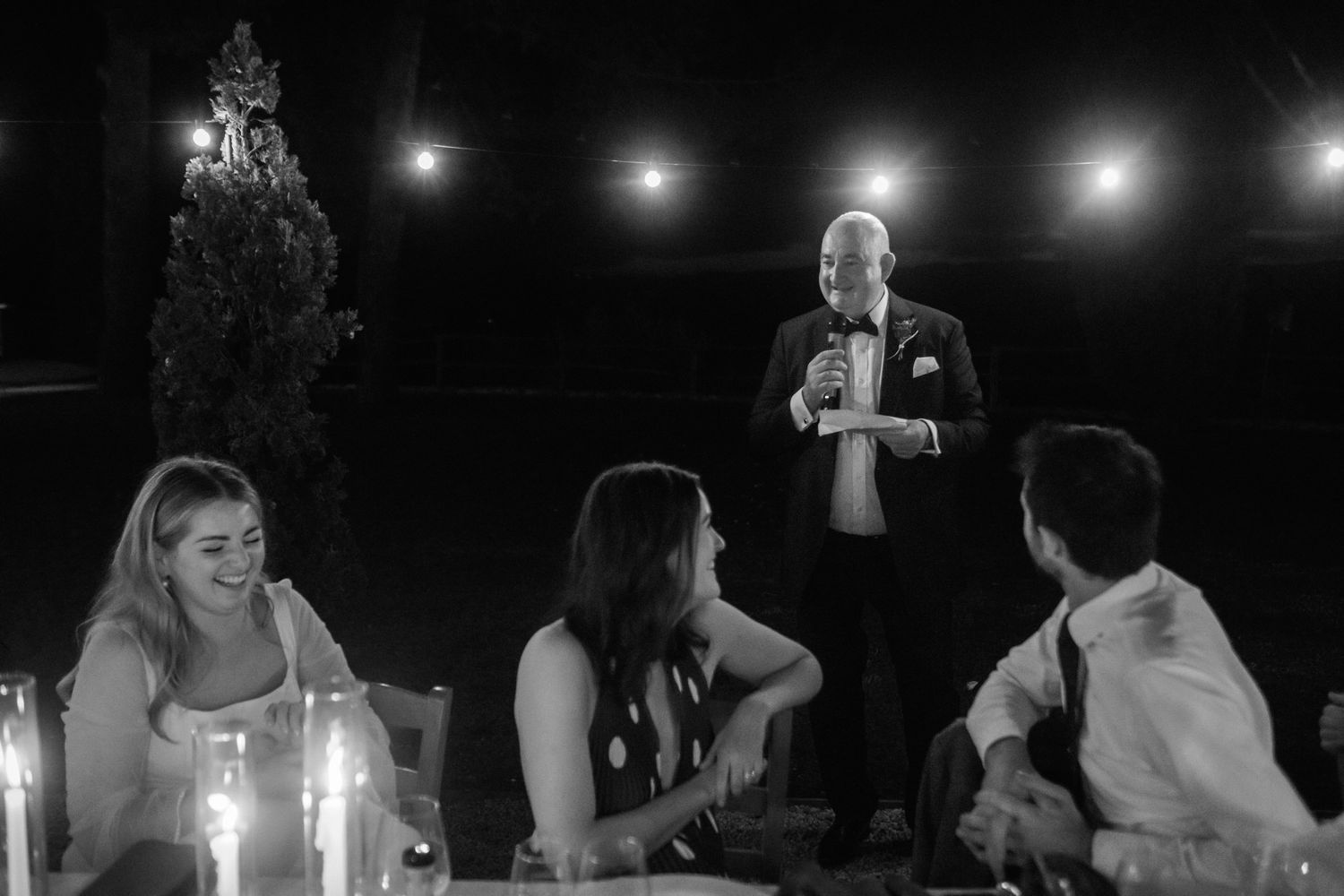Black and white photo of a man in a bow tie giving a toast while guests smile and watch at a candlelit table under string lights.