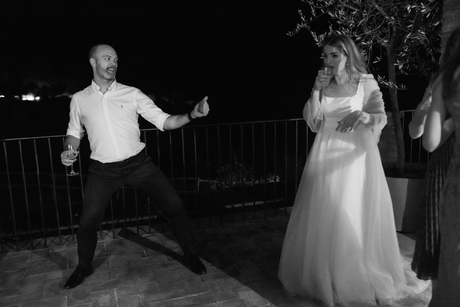 Black and white photo of a groom dancing playfully on a terrace at night while the bride in a flowing gown watches and holds a drink.