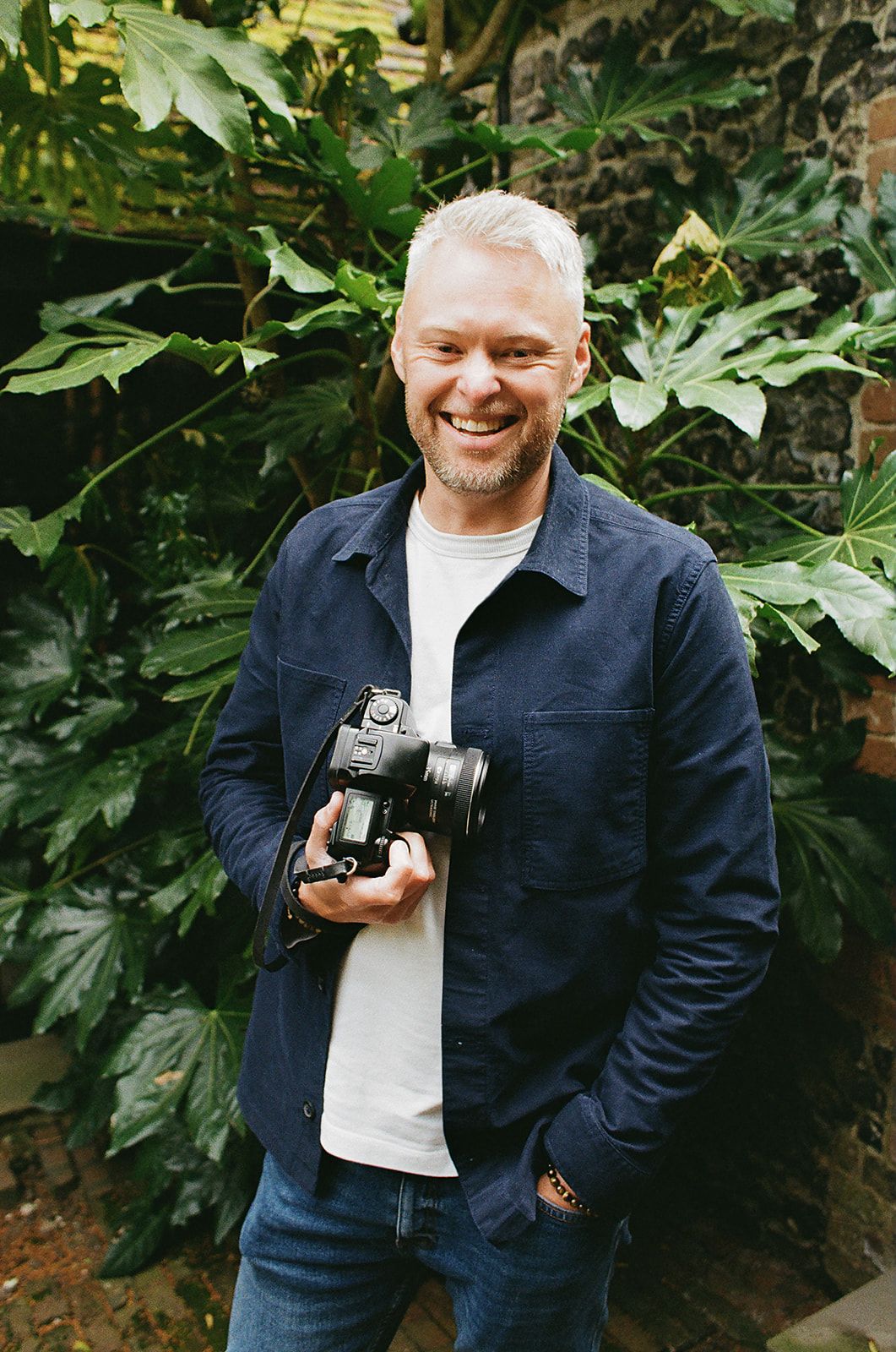 Photographer in navy jacket holding vintage camera against lush green foliage background.