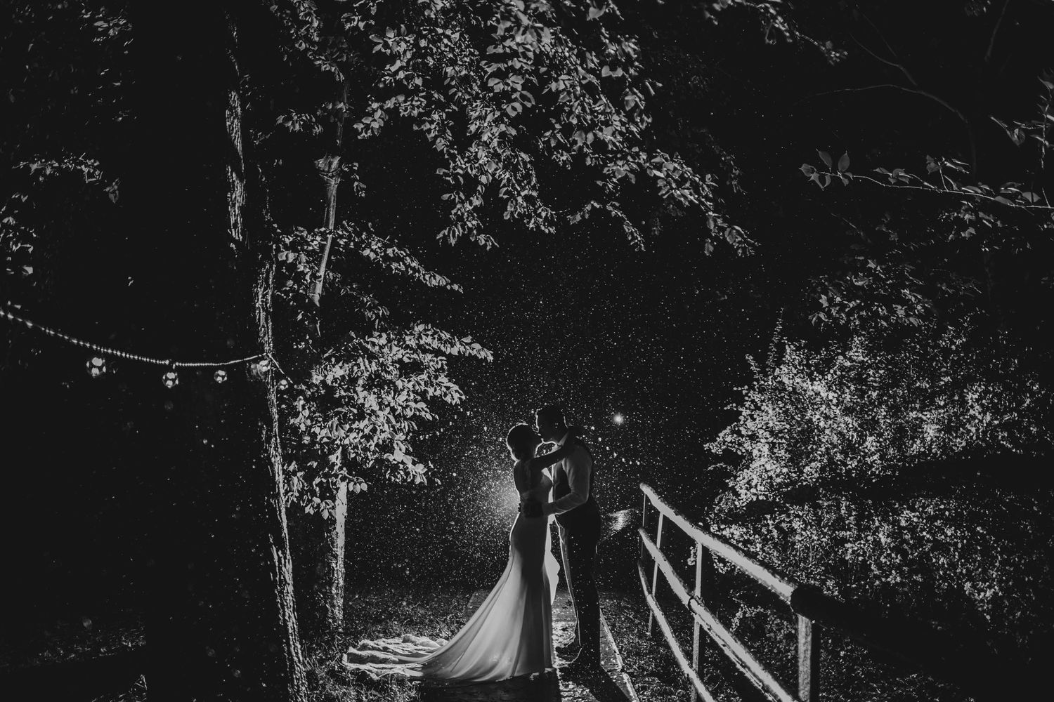 A dramatic black and white nighttime wedding portrait on a wooden bridge surrounded by trees.