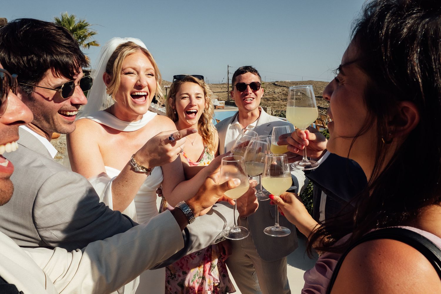 Wedding guests laughing and toasting with champagne glasses outdoors on a sunny day.