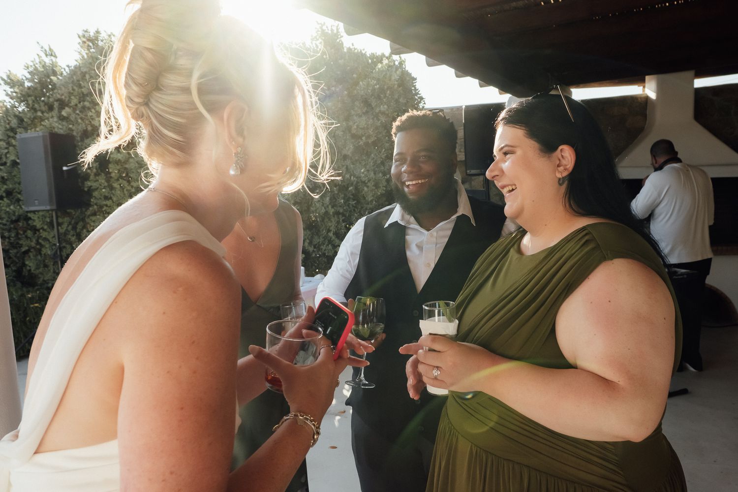 Guests socializing at a wedding reception, holding drinks outdoors in warm sunlight.