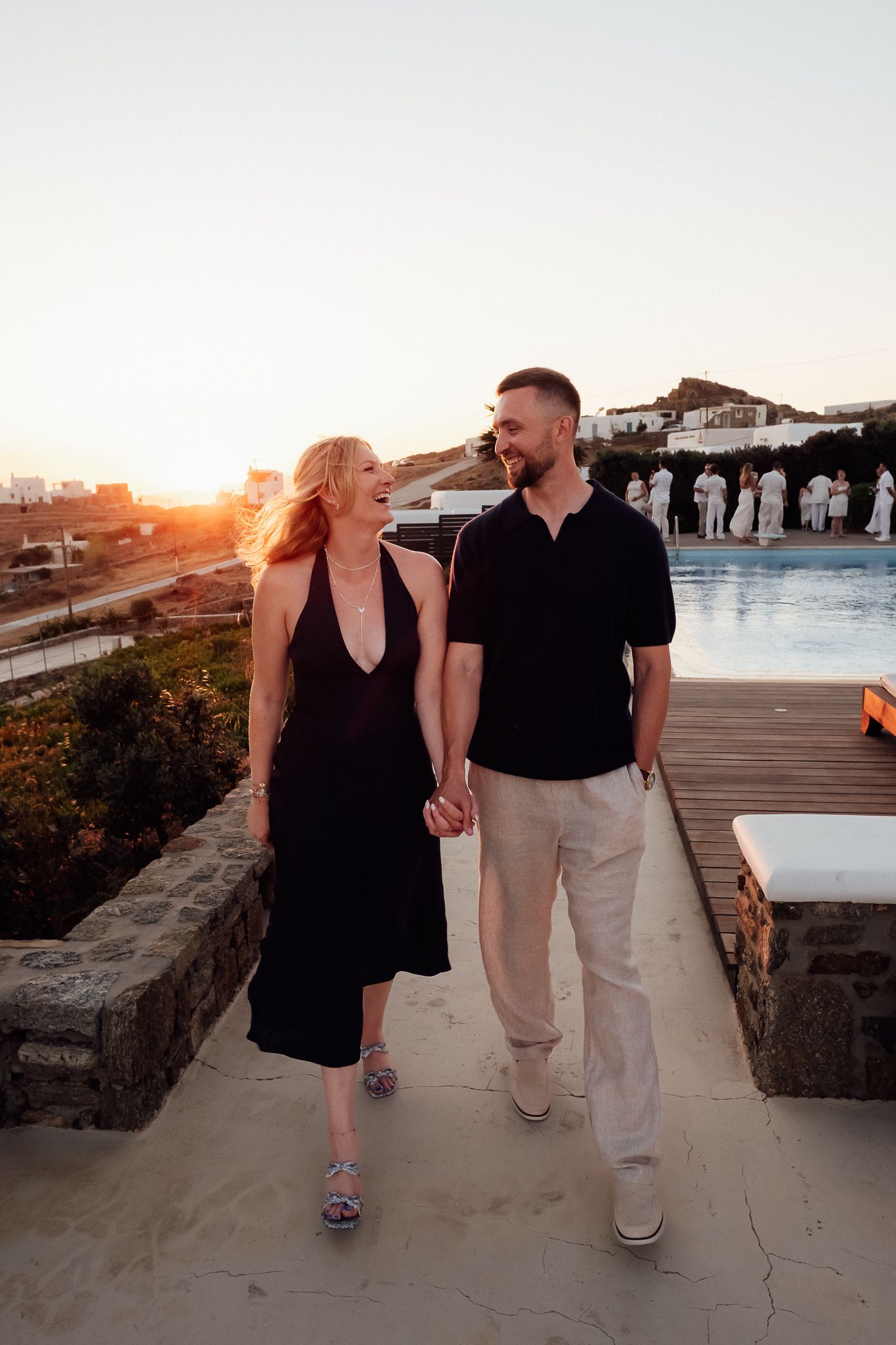 Couple holding hands walking by a pool at sunset, woman in black dress, man in black shirt and beige pants.