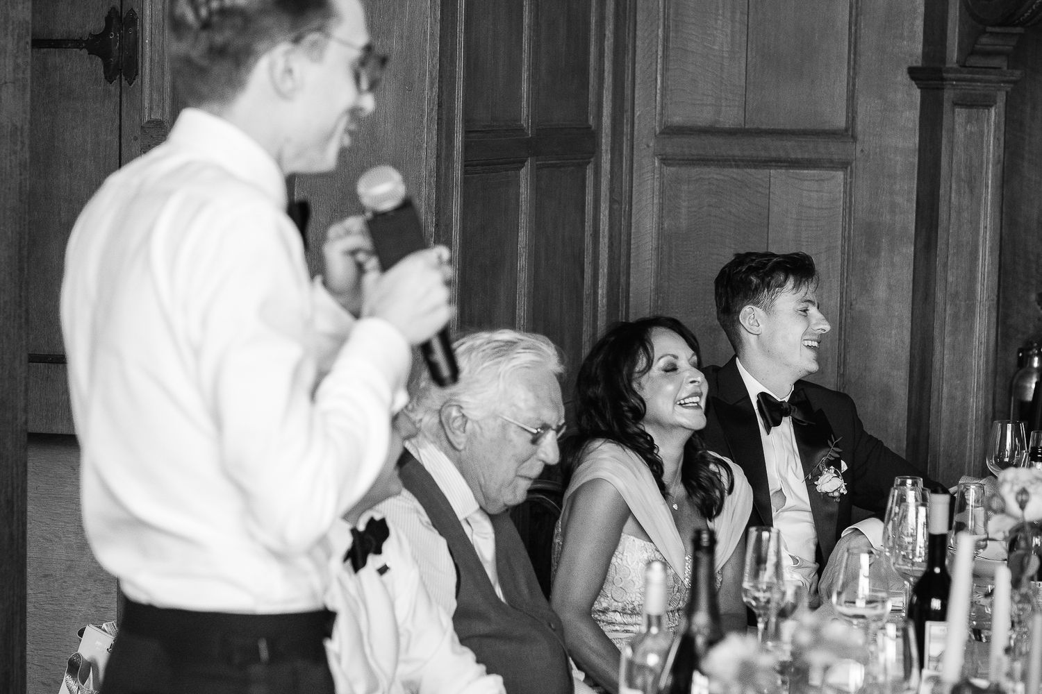 Black and white photo of wedding guests laughing at head table during speech.