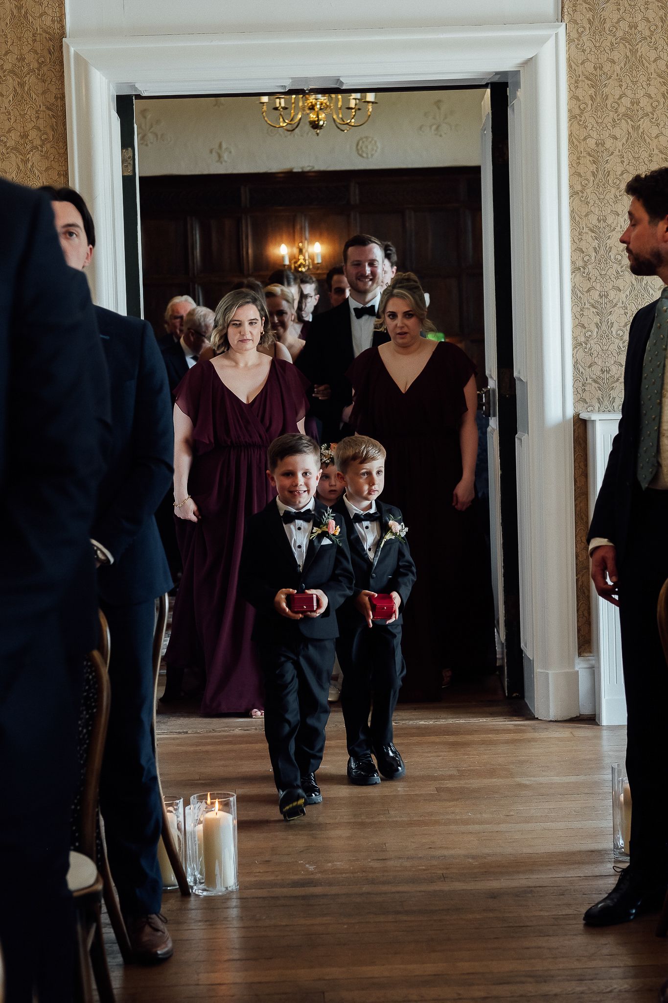 Two young ring bearers in tuxedos walk down the aisle at an elegant wedding ceremony with guests looking on.