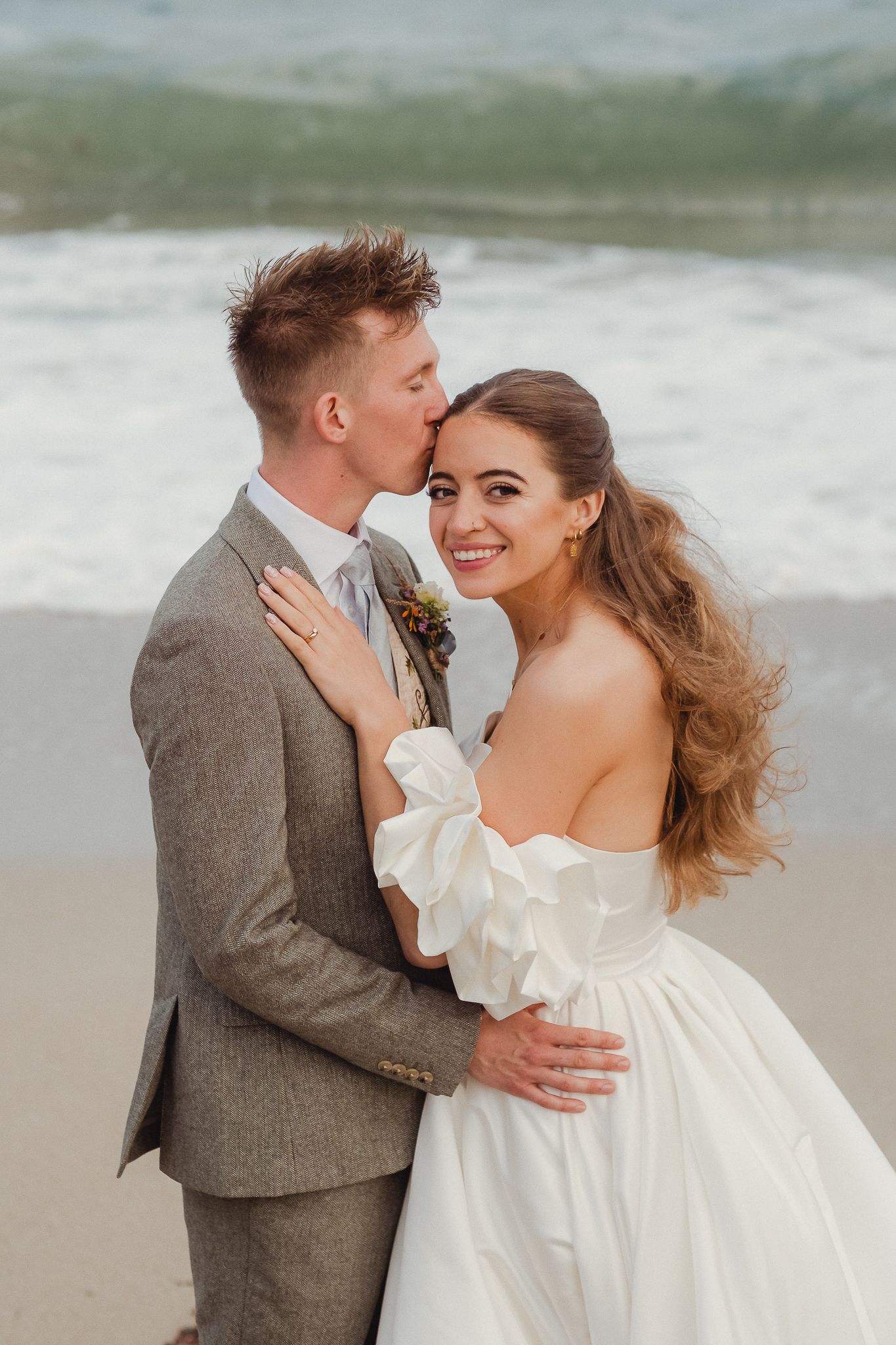 A romantic beach wedding moment with a couple embracing, bride in off-shoulder white gown and groom in gray suit.