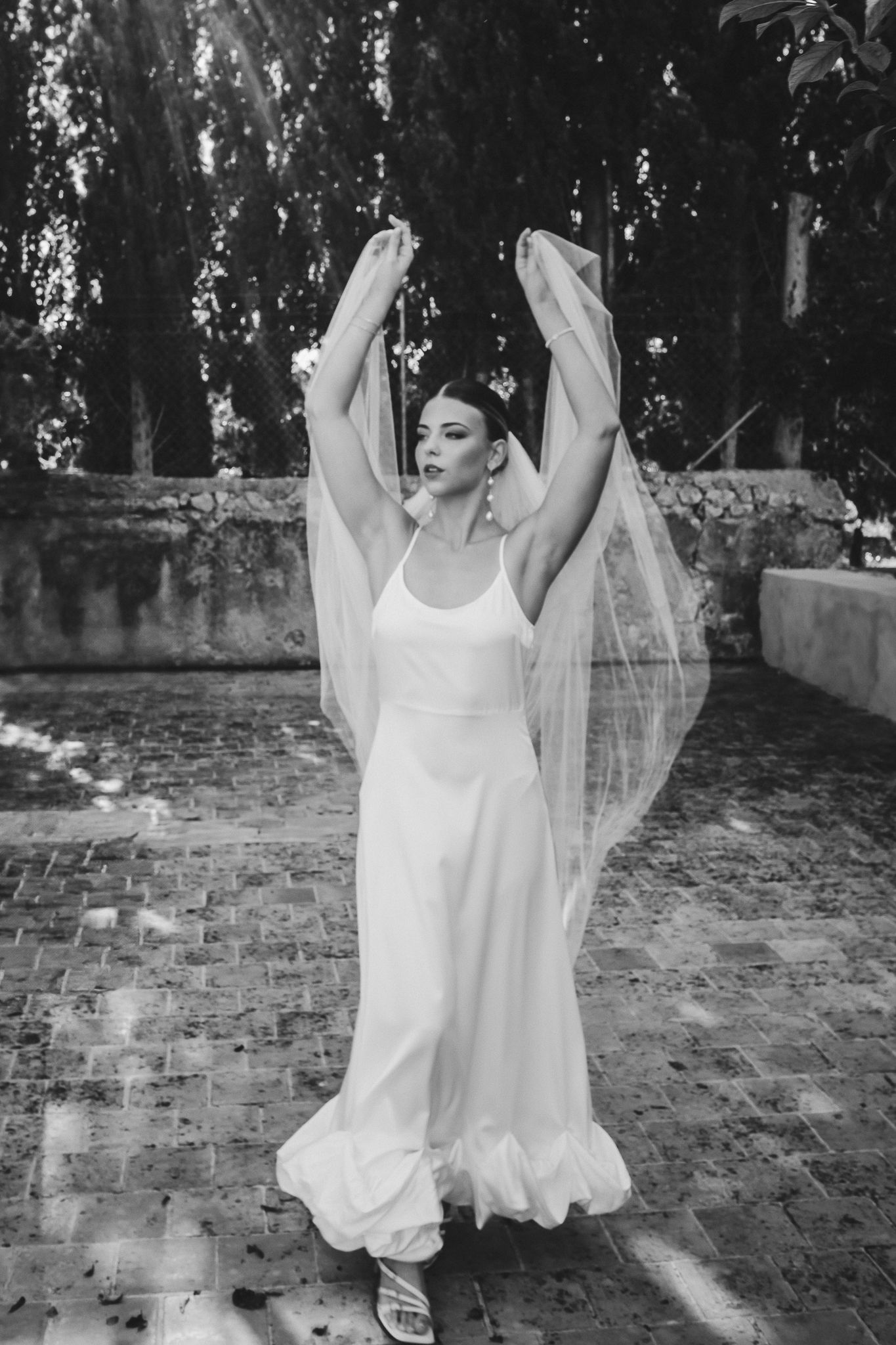 A bride in a flowing white dress and veil poses gracefully with raised arms on a brick patio surrounded by trees.