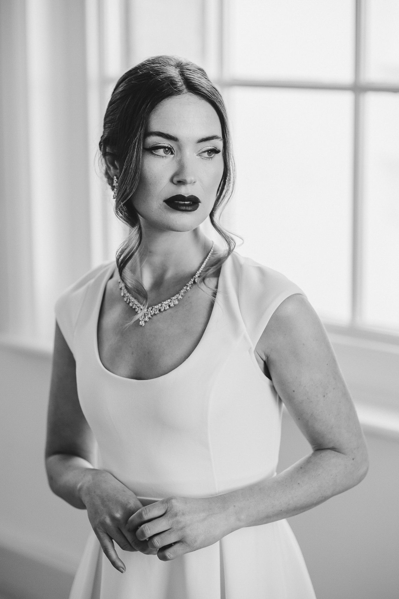 Black and white portrait of a woman in a white dress wearing dark lipstick and a necklace by a window.