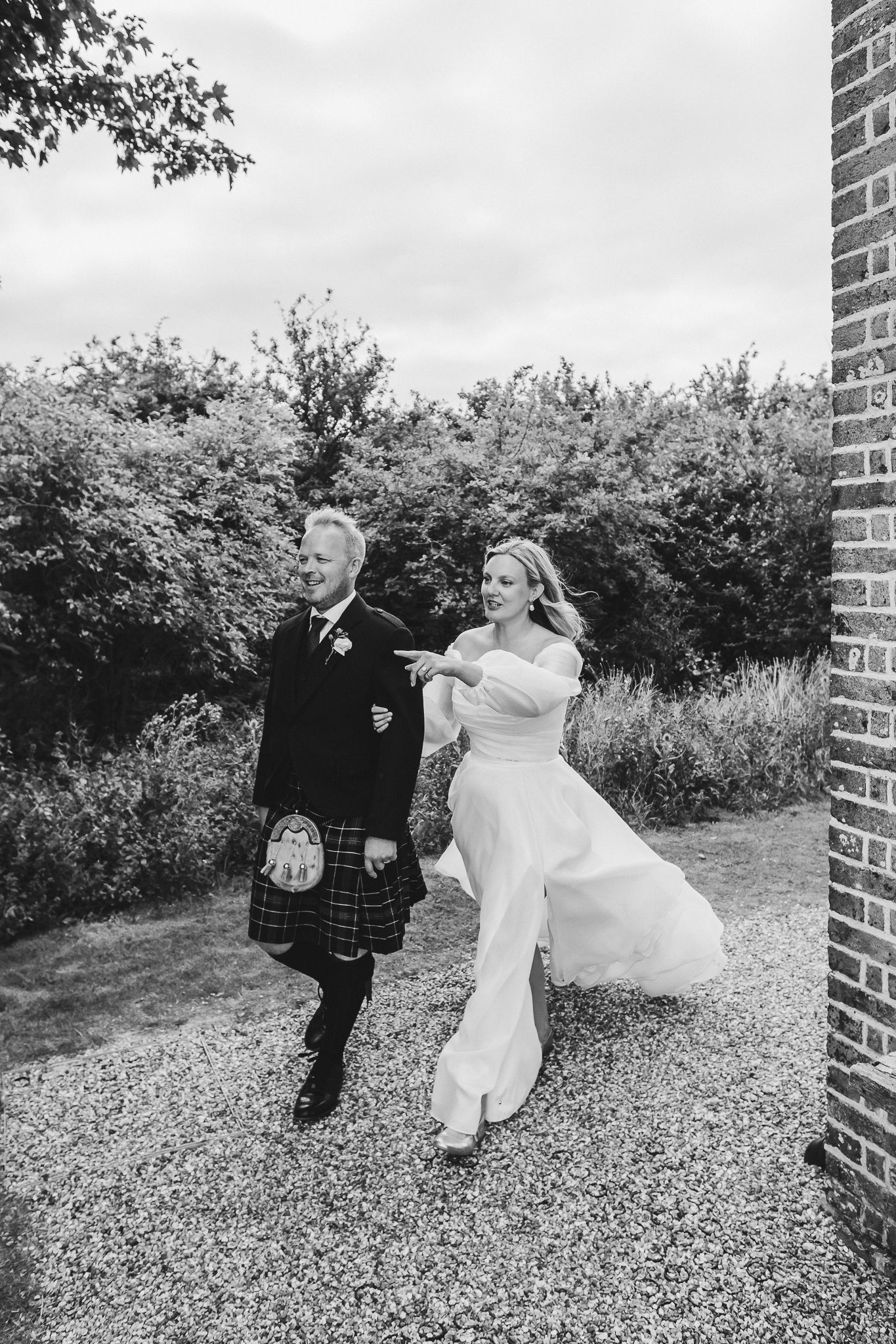 A black and white wedding photo showing a couple walking together on a gravel path, with the groom in a traditional kilt.