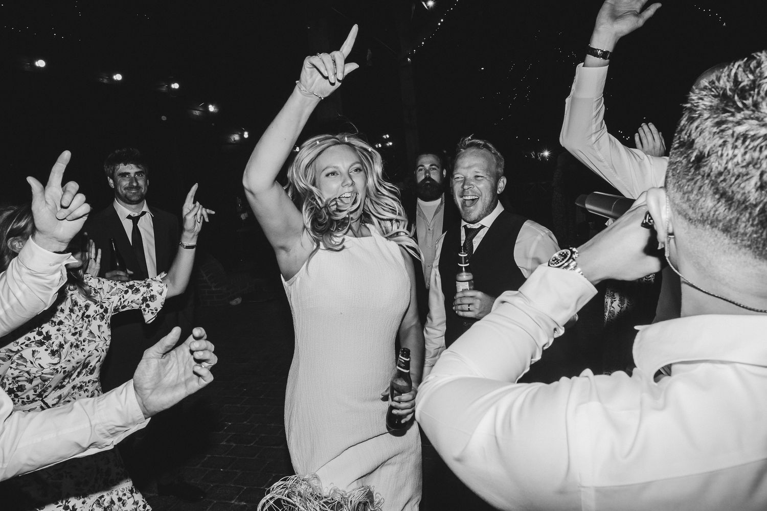 Black and white photo showing wedding guests dancing and celebrating with raised arms on a dance floor.