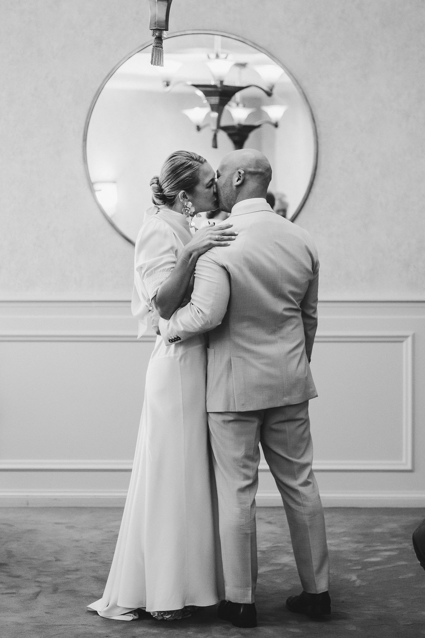 A couple shares an intimate moment during their wedding, captured in black and white against a simple backdrop with a round mirror.