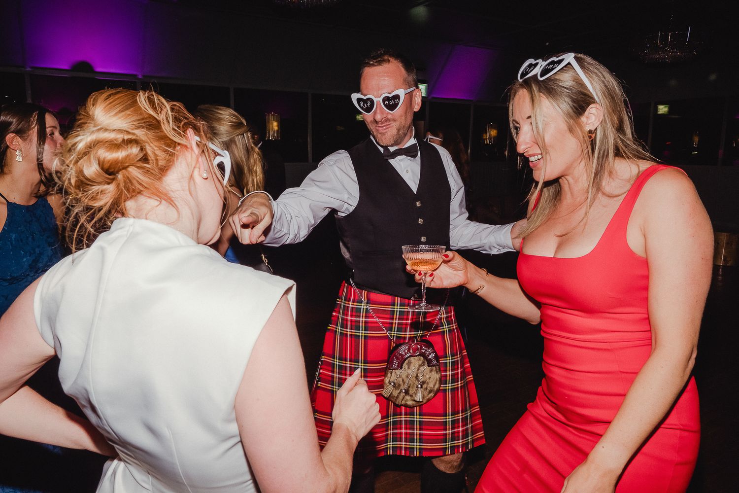 Party guests dance and socialize at a lively event with purple lighting and someone wearing a traditional kilt.