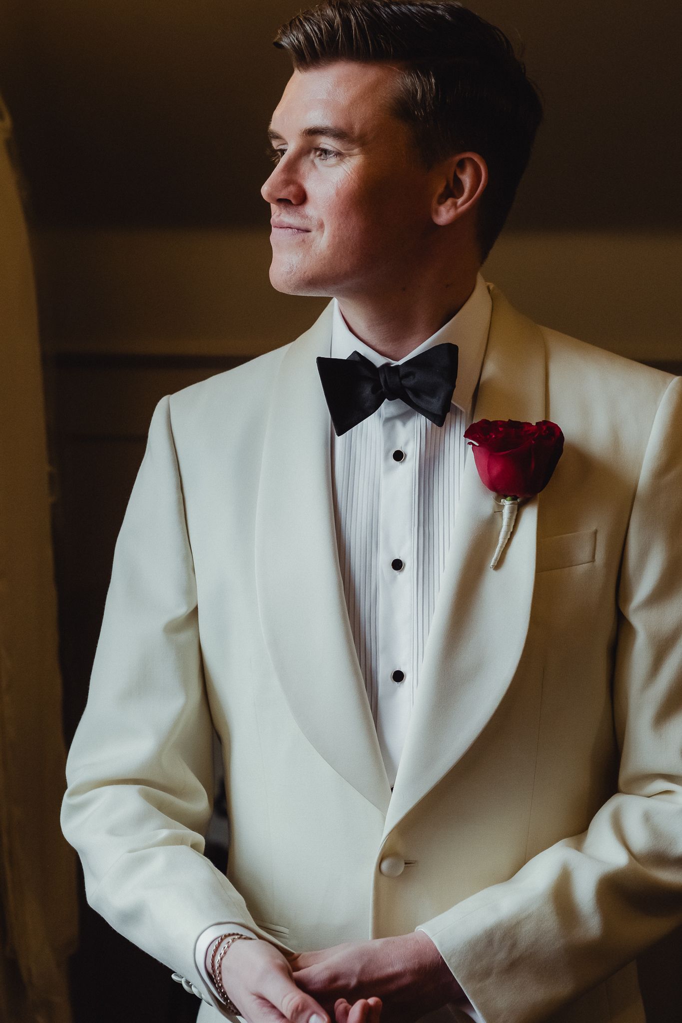 A groom in a classic white tuxedo jacket with black bow tie gazes thoughtfully to the side in a formal portrait series.