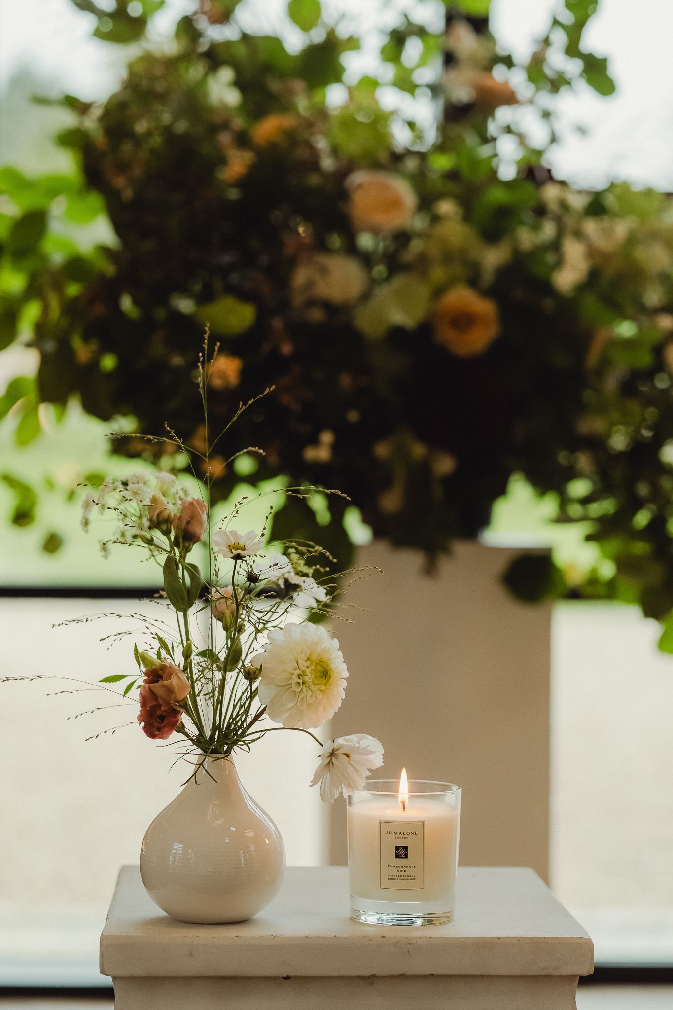 Minimalist white vase with delicate flowers and lit candle arranged on light surface against window with natural light.