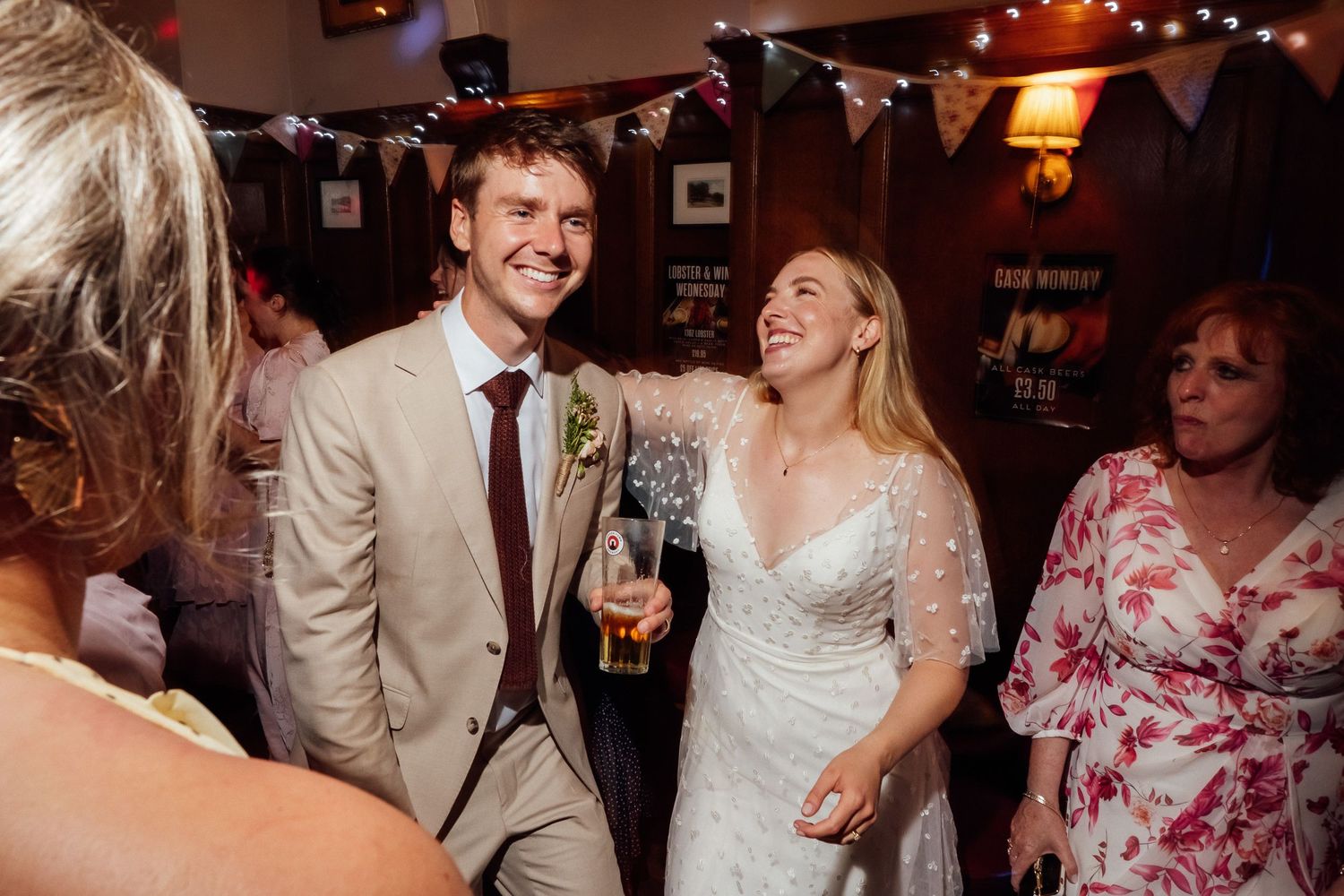 Smiling groom in a tan suit and laughing bride in a white lace dress celebrate with guests at a lively pub wedding reception.