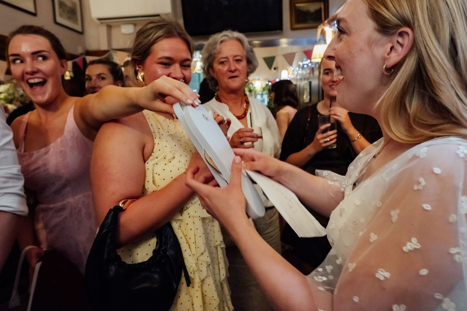 Smiling women in a lively crowd gather around a woman in a white floral top who holds out papers in a warm, festive indoor setting.