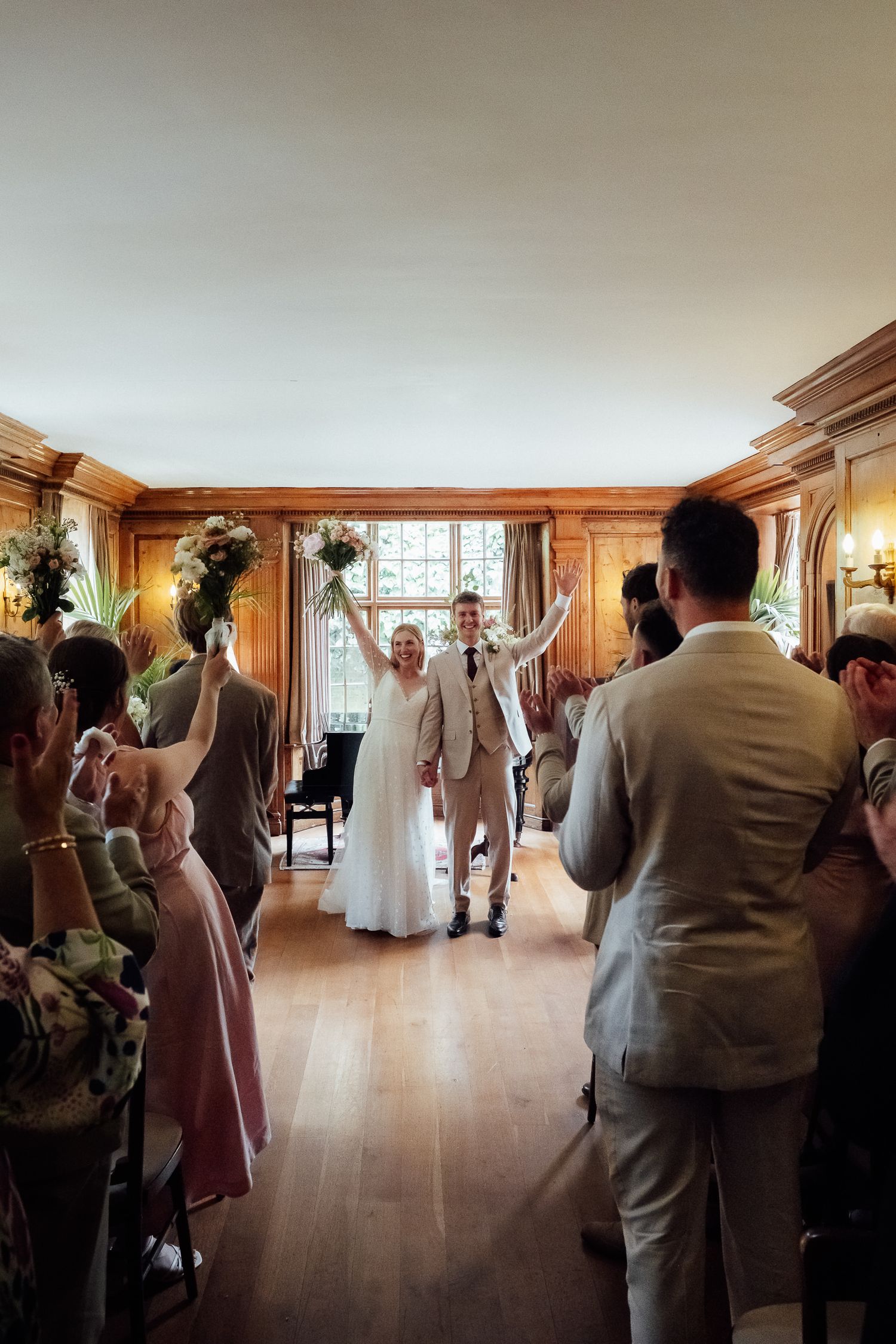 Newlywed couple in a wedding dress and tan suit raise their arms in celebration while guests cheer in a wood-paneled room.