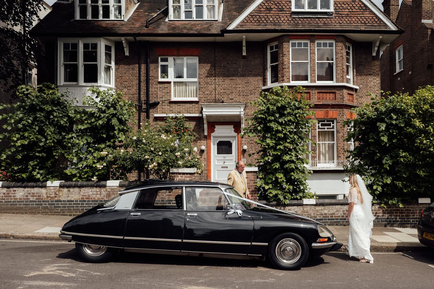 A bride in a white gown stands beside a vintage black Citroën DS parked outside a red brick townhouse with green hedges.