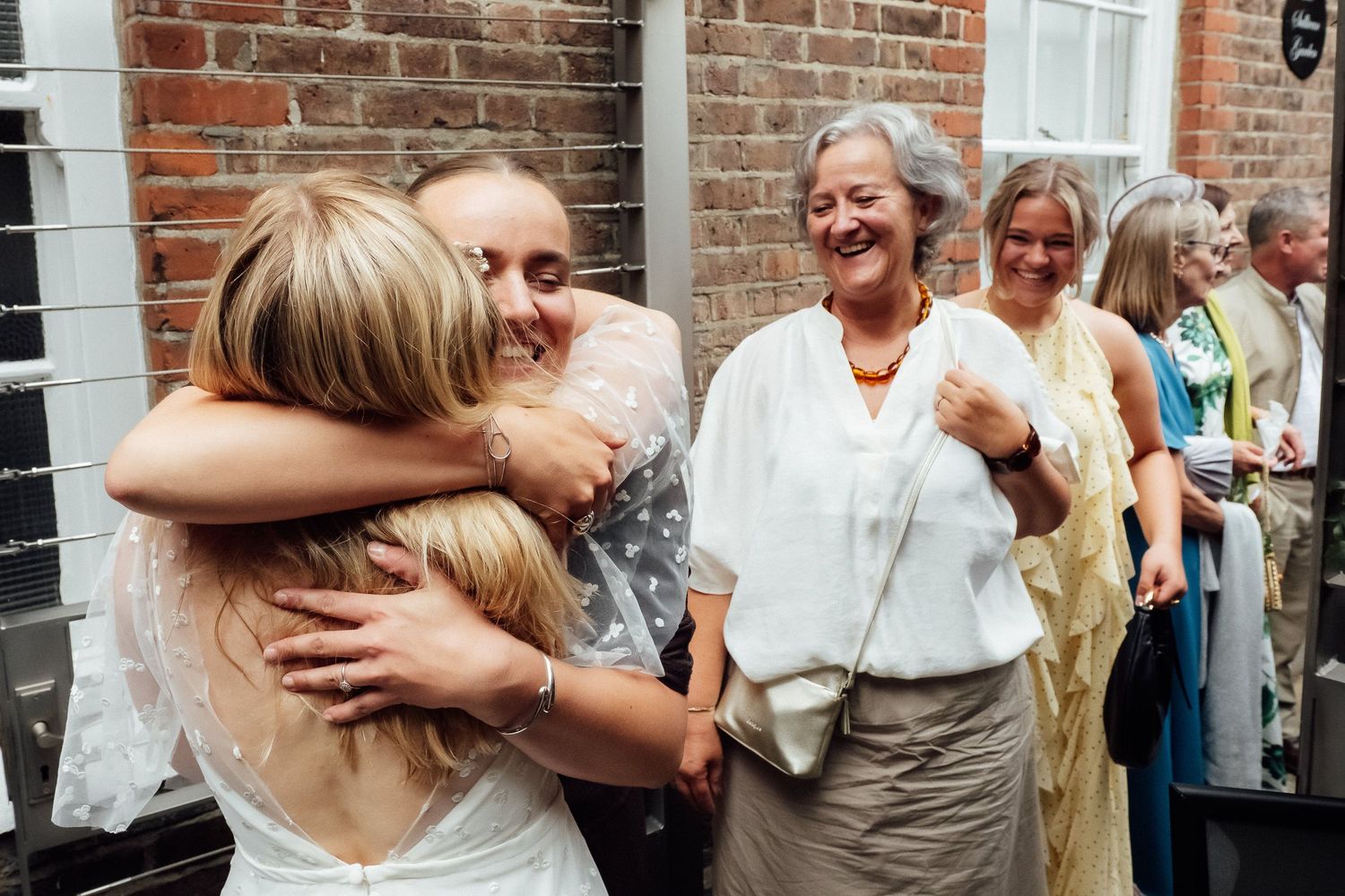 Smiling guests in summer dresses embrace and celebrate outside a brick building at a joyful wedding gathering.