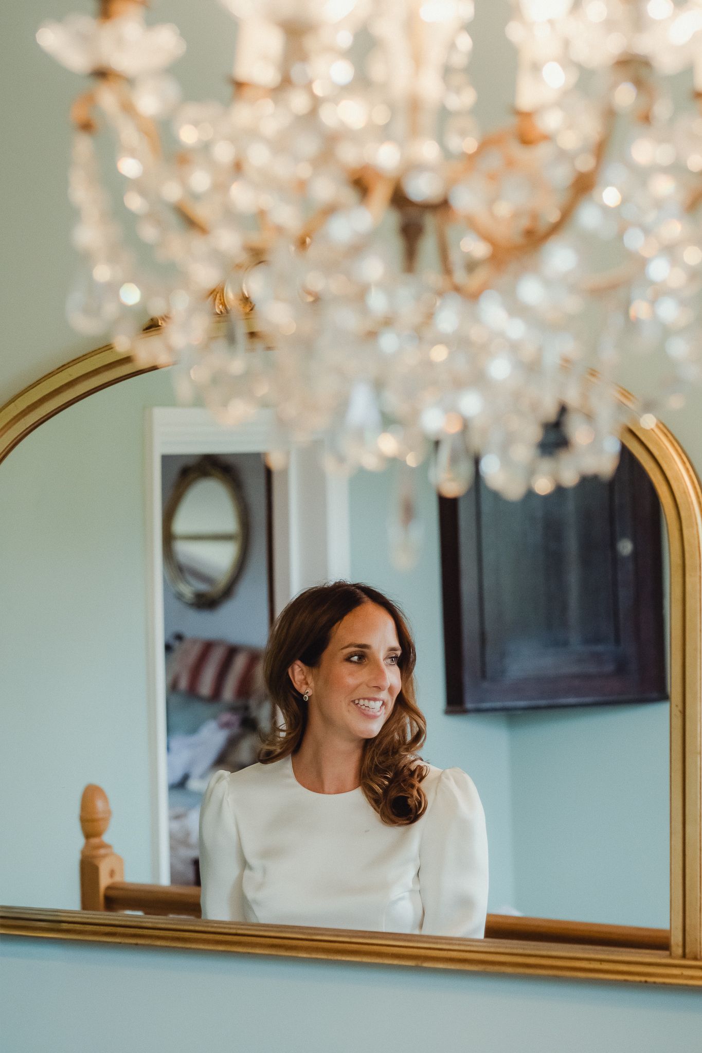 Elegant reflection in gold-framed mirror under crystal chandelier in mint green room with soft natural lighting.