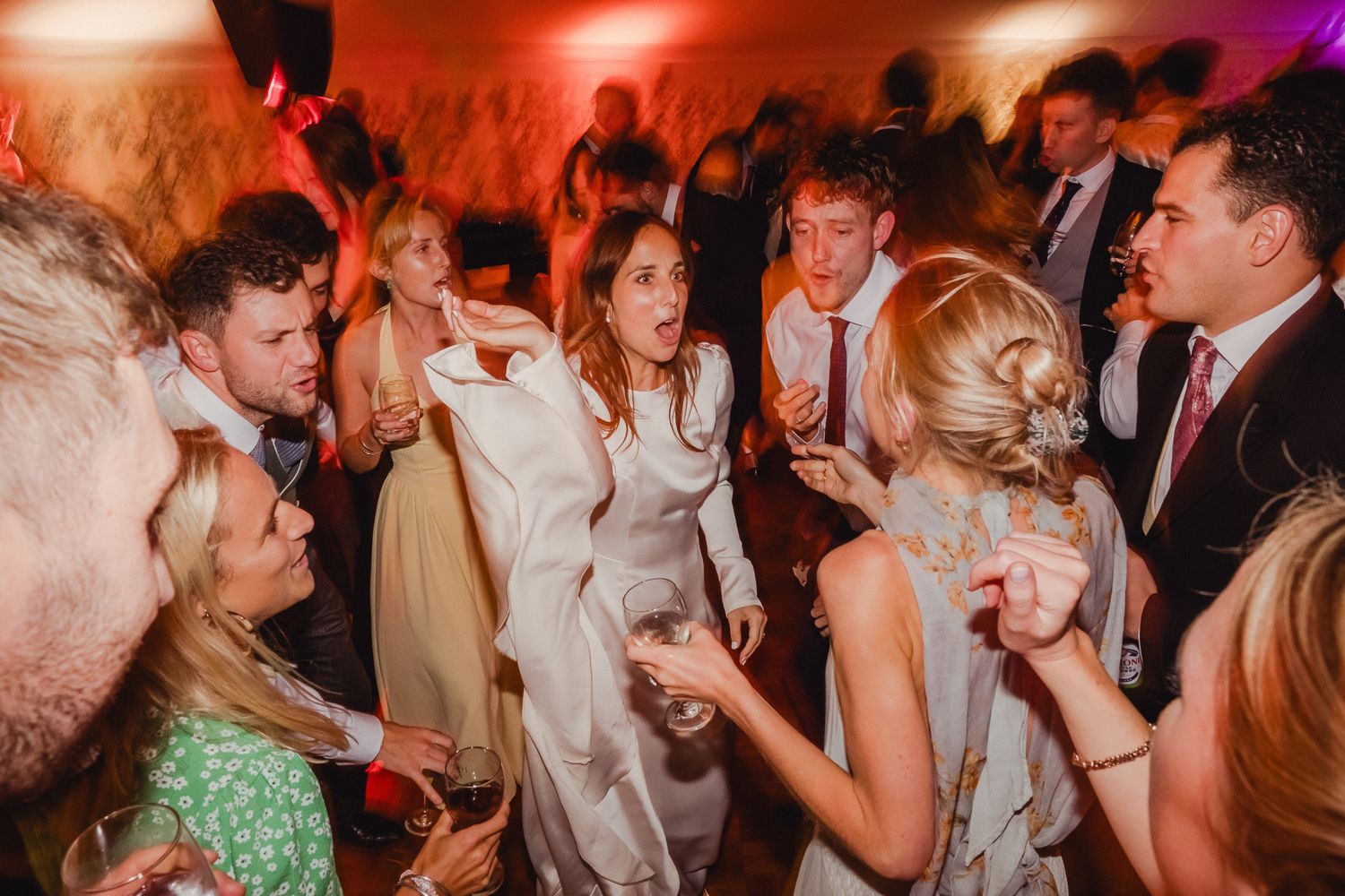 Wedding guests dancing and celebrating on a crowded dance floor illuminated by red and purple lights.
