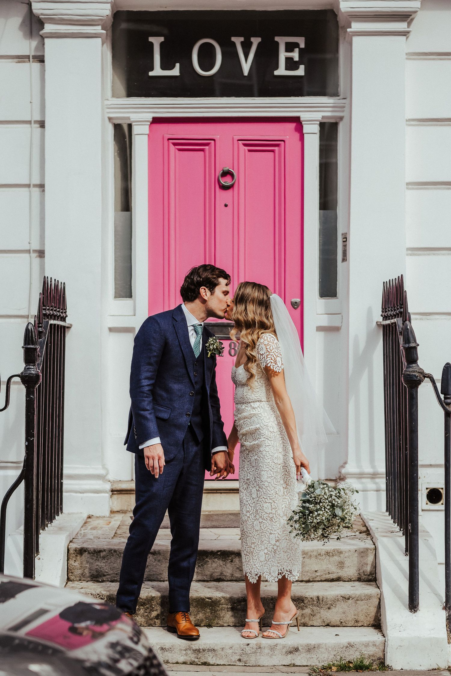 Wedding couple sharing a romantic kiss on steps in front of pink door and LOVE sign in urban setting.