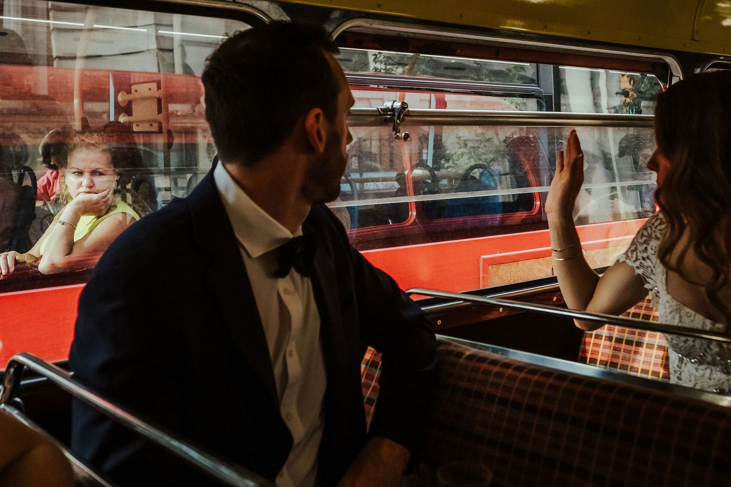 Elegantly dressed couple sits in vintage red double-decker bus while onlookers wave through window.