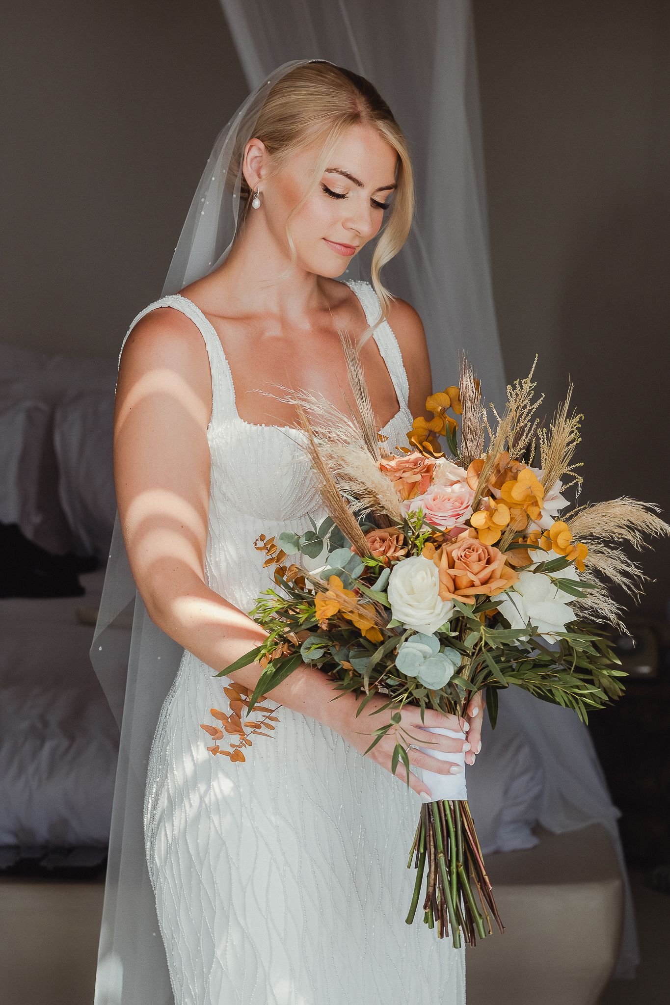 A bride in a white gown admires her stunning autumn-themed bouquet with orange lilies and dried wheat stems.