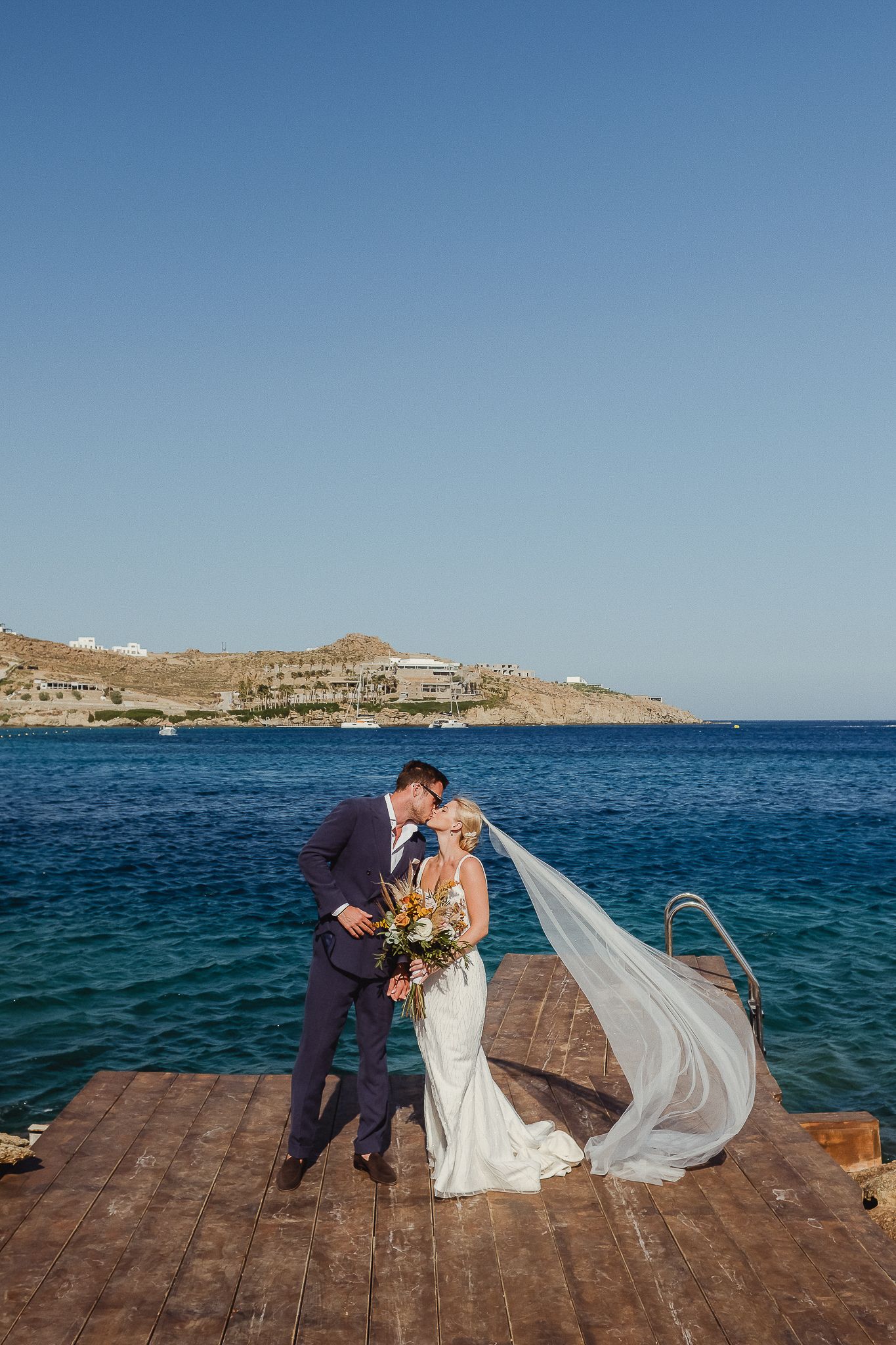 A romantic wedding moment on a wooden dock overlooking turquoise Mediterranean waters and coastal cliffs.