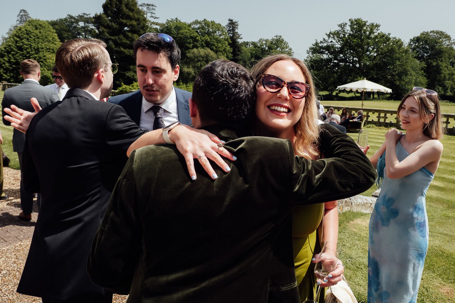 Guests hugging and socializing at an outdoor wedding reception on a sunny day in a garden setting.