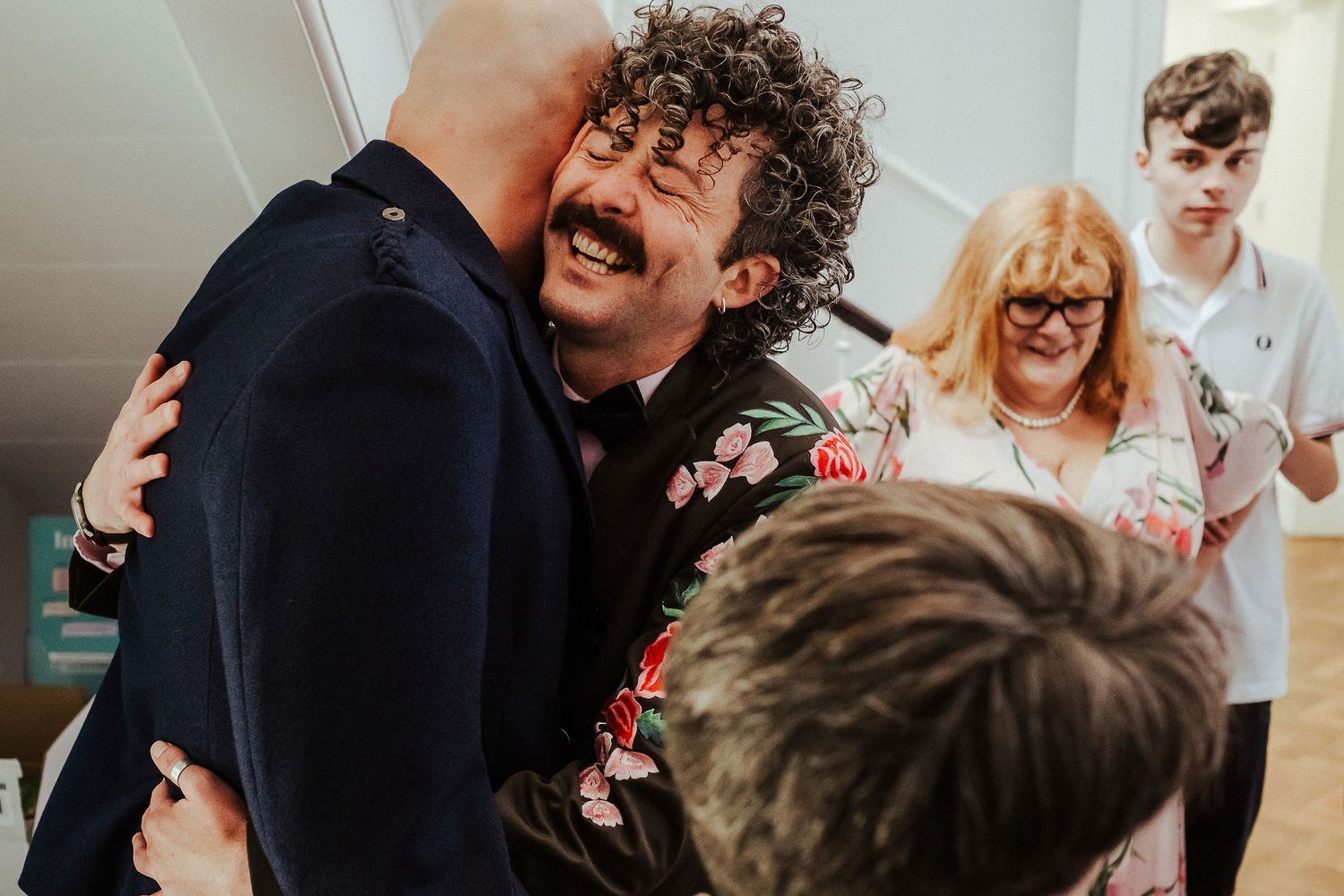 A laughing curly-haired man in a floral jacket hugs a bald man in a dark suit while guests smile in the background.