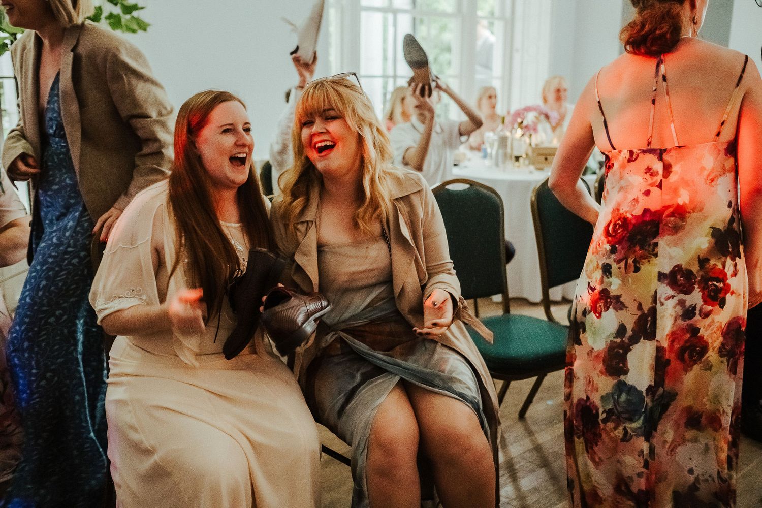 Two women laugh joyfully while seated together at a wedding reception, surrounded by guests in elegant floral dresses.