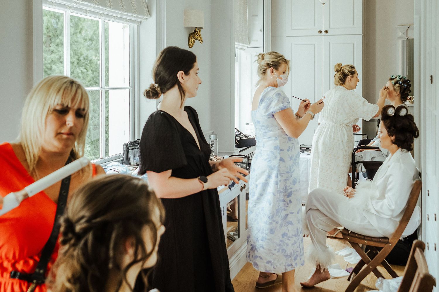 Wedding preparation scene showing bridesmaids helping with bridal preparations in a sunlit room.