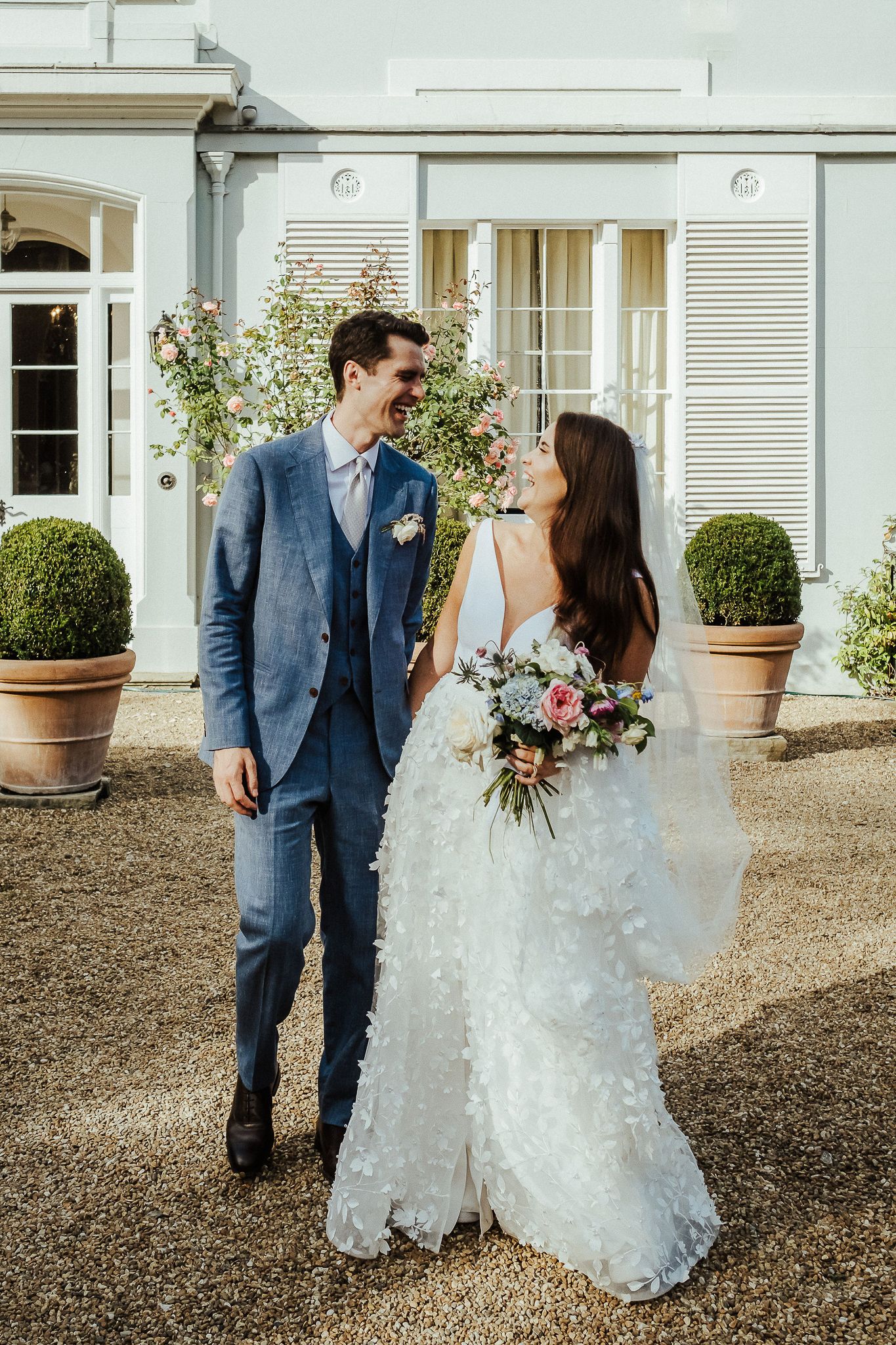 Elegant couple poses outside a white mansion with potted boxwoods and climbing vines.